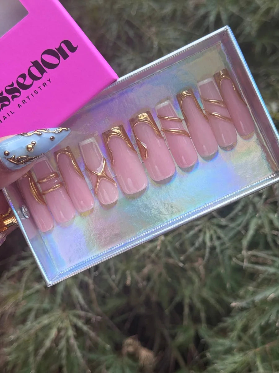 Set of pink medium press-on nails with glossy finish and gold line art designs. The classic-shaped nails feature curved and criss-cross patterns in gold metallic.