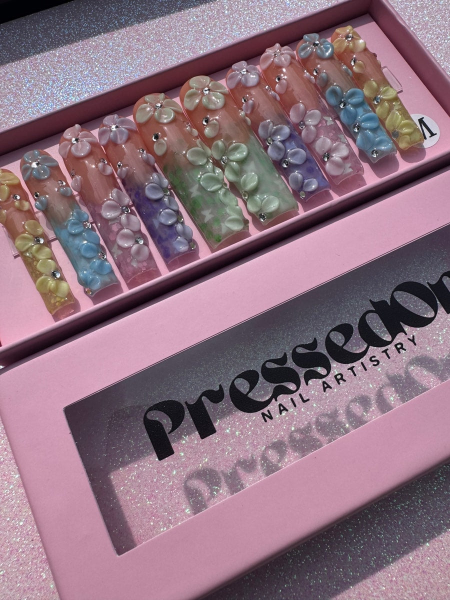 Extra-long press-on nails in pastel yellow, blue, pink, green, and purple with a floral design and rhinestone accents.