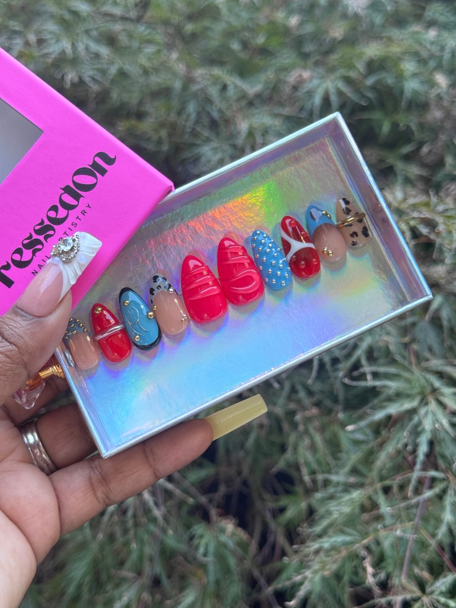 A set of short press-on nails with varying designs, including solid red and blue, polka dots, leopard print, and gold embellishments.