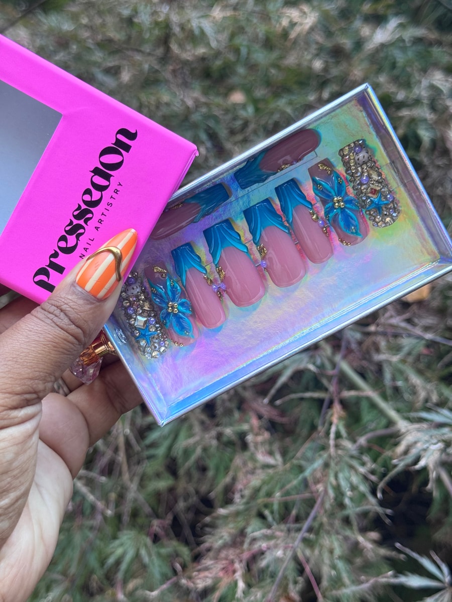 Medium-length pink press-on nails with sculpted blue tips, blue floral charms, and iridescent rhinestone accents.