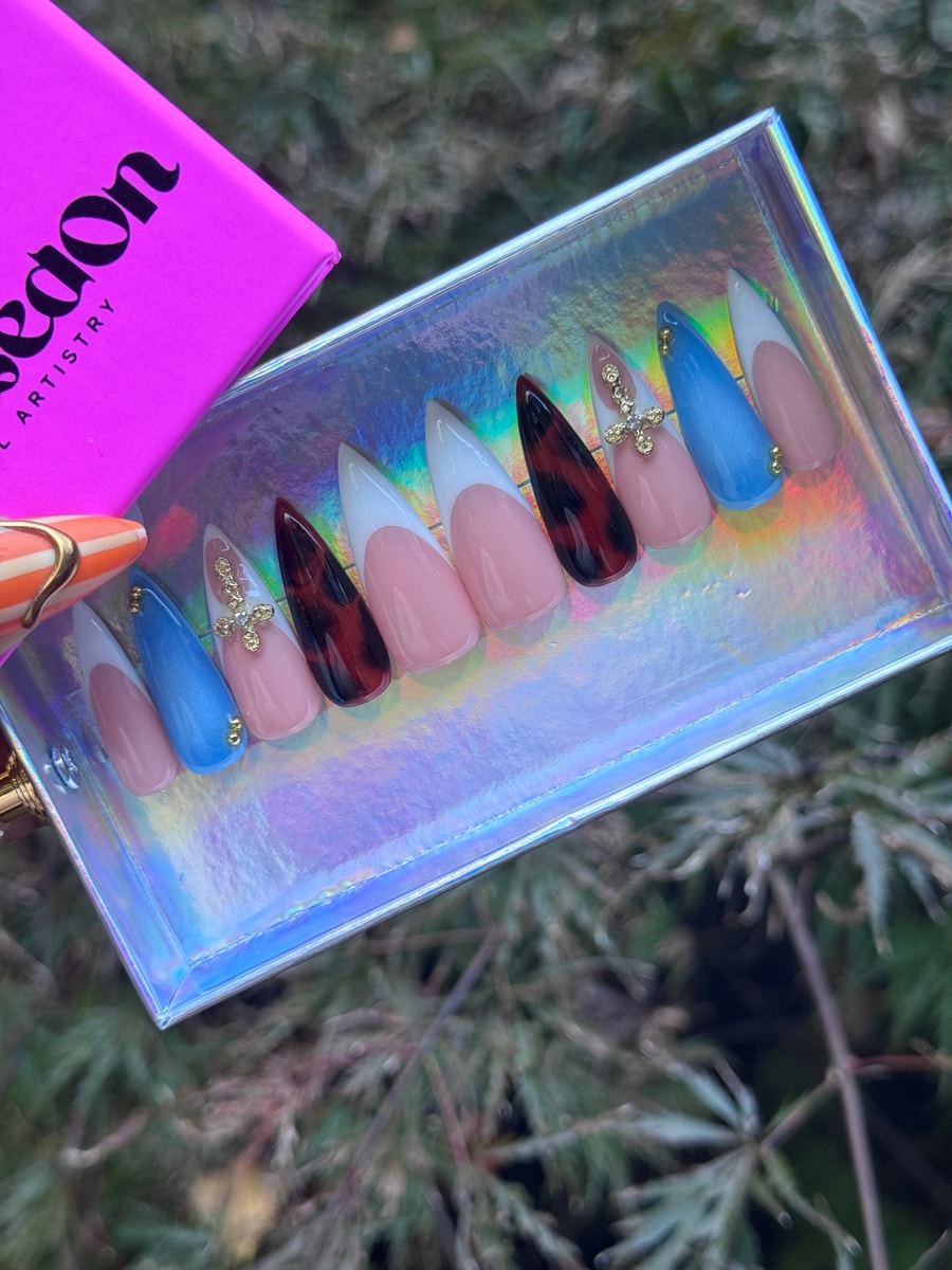 Set of stiletto press-on nails with French twist designs. Colors include pink, blue, white and tortoise shell. Some nails feature gold cross charms and beads.