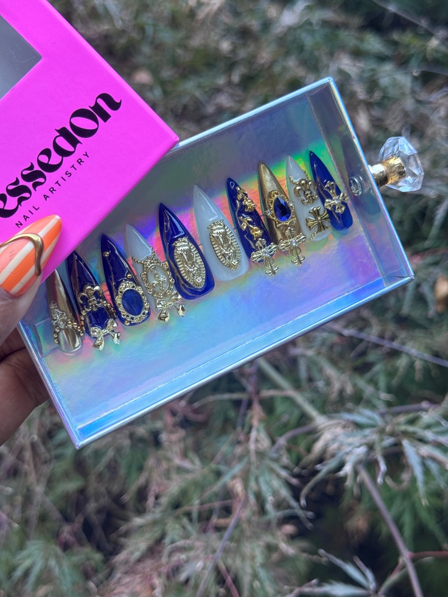 Stiletto shaped press-on nails in blue, gold, and white with religious and royal themed gold charms, including crosses and emblems.