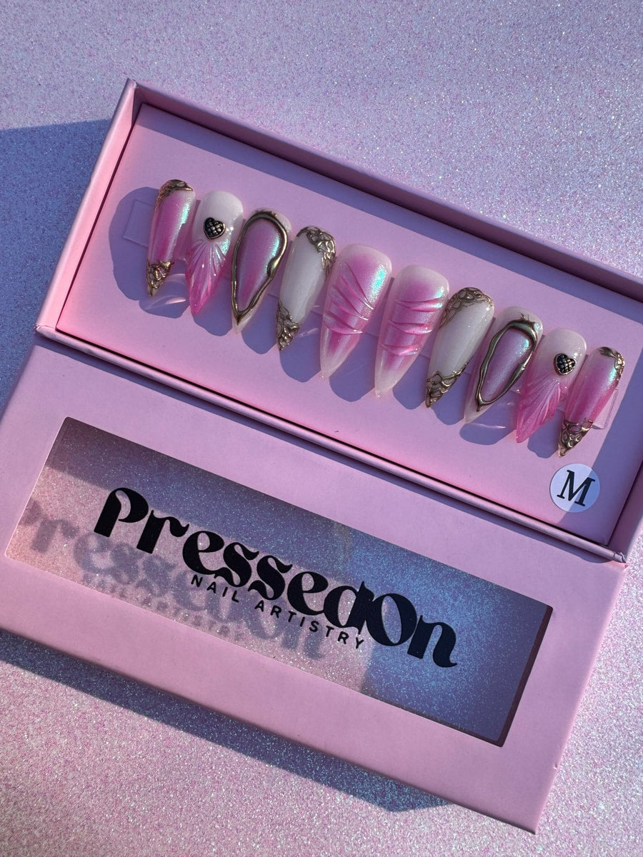 Set of press-on nails features pink paradise designs with gold details. Nail designs include iridescent pink, solid white, and unique embellishments.