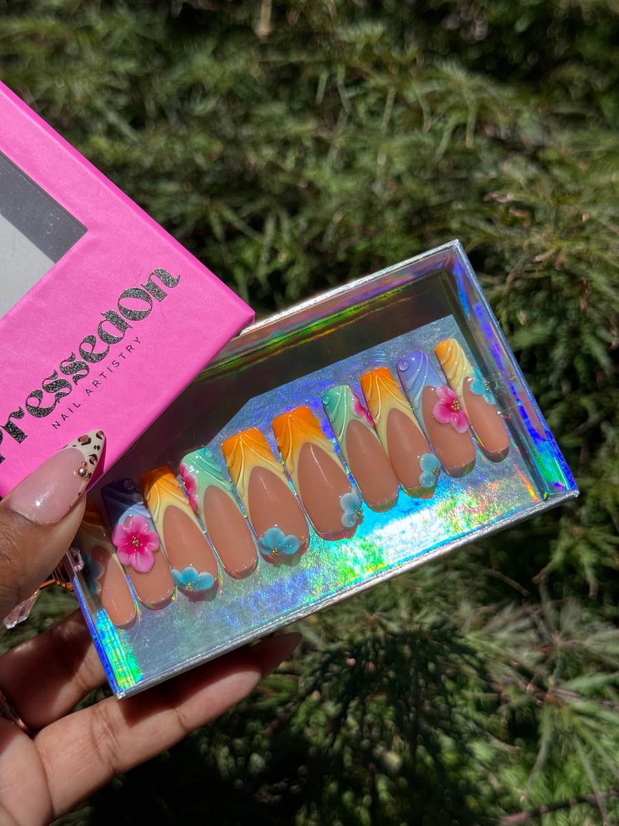 Medium-length press-on nails with nude base and multi-colored French tips in purple, blue, green, yellow, and orange, accented with delicate 3D flower decorations.