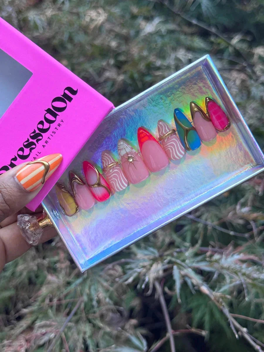 Short almond press-on nails with abstract designs. The nails feature neon colors, white wavy lines, gold accents, and glitter details.