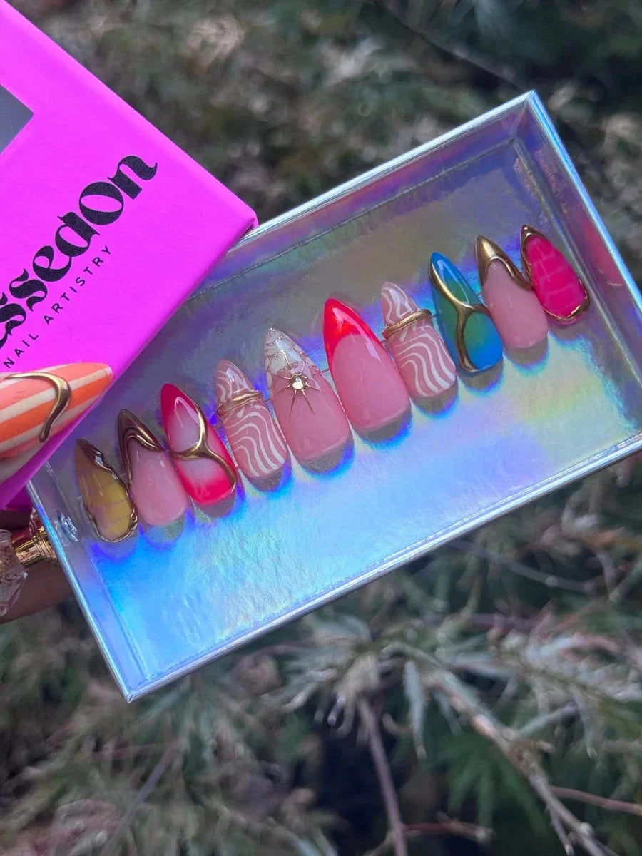 Short almond press-on nails with abstract designs, including neon colors, pink base coats, gold accents, and white swirl patterns.