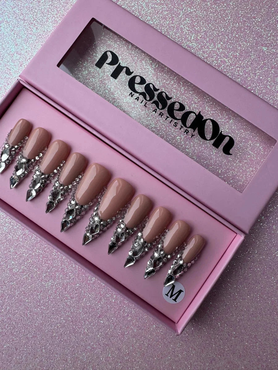 Nude long stiletto press-on nails with a cascading design of clear rhinestones and sparkling crystals. The jewels accent the base of each nail, providing a bling effect.