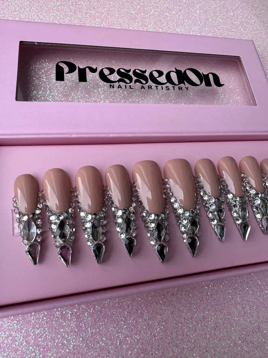 Nude stiletto press-on nails feature clear and black rhinestones cascading down from the cuticle. The nails are long and pointed, creating a bling effect.