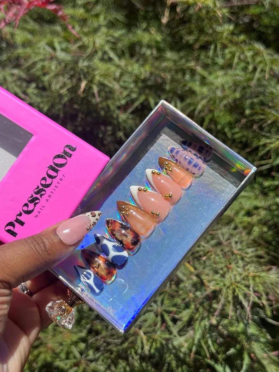 A set of short press-on nails with a nude base and cheetah print accents. Some nails feature a white tip and a blue and white cow print design. Gold studs adorn some of the nails.