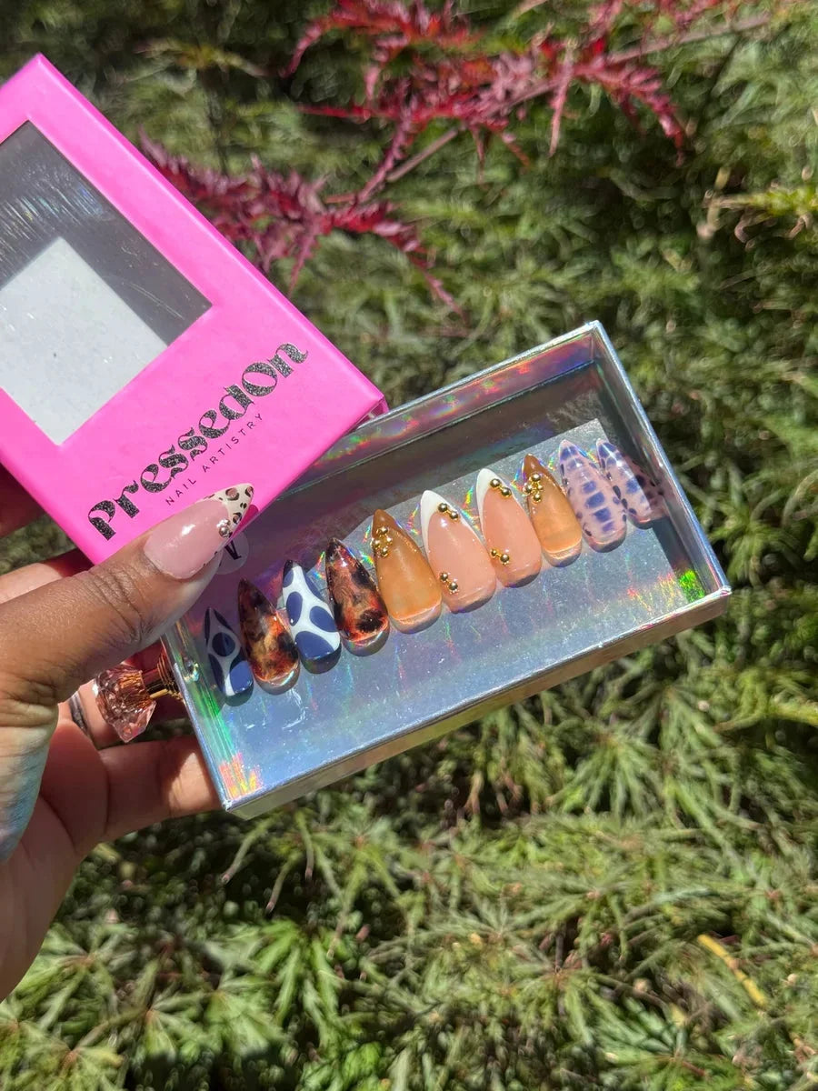 Short, almond-shaped press-on nails with cheetah print, cow print, and nude designs, some accented with gold studs.