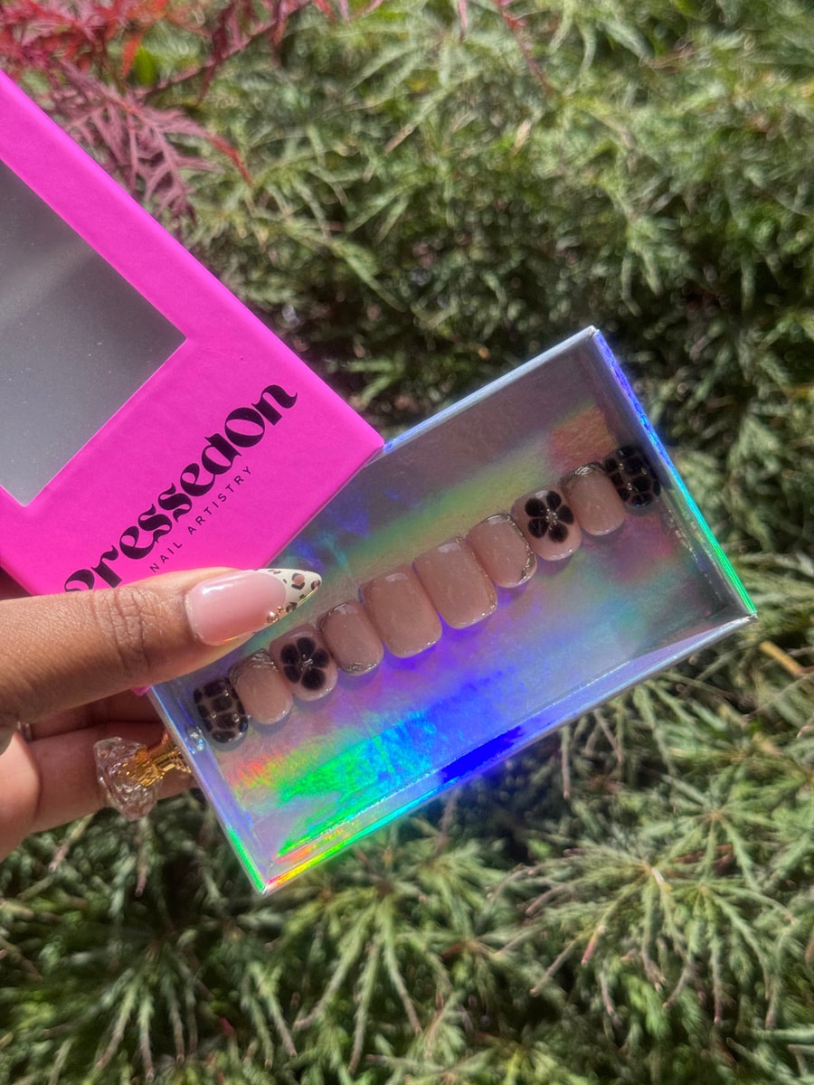 These are short, nude-colored press-on nails adorned with delicate black flower accents. A touch of glitter complements the floral design on some nails.