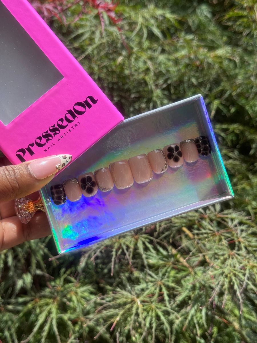 Nude press-on nails feature black flower designs and checkered detailing, along with small silver accents.