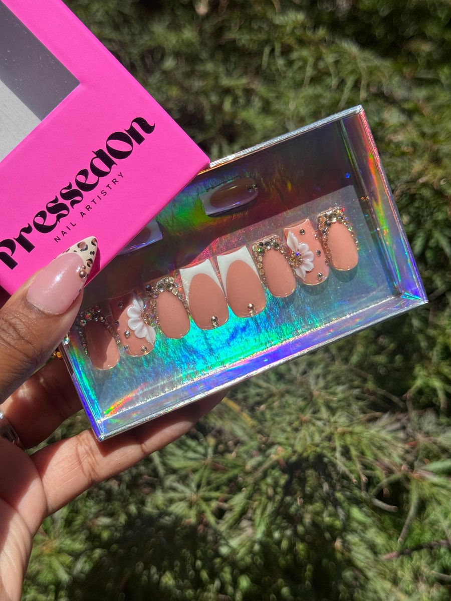 These are short, nude press-on nails. Several nails have white French tips and rhinestone accents, while two nails feature 3D white flower charms and gold studs.