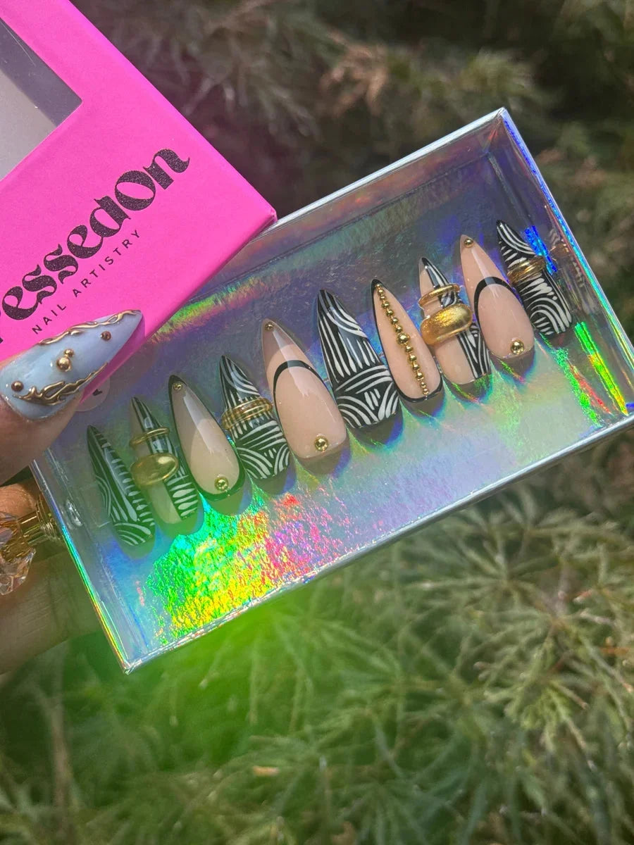 Set of stiletto press-on nails with a nude base and black and white patterns. Nails feature zebra print, gold rings, and gold bead accents.
