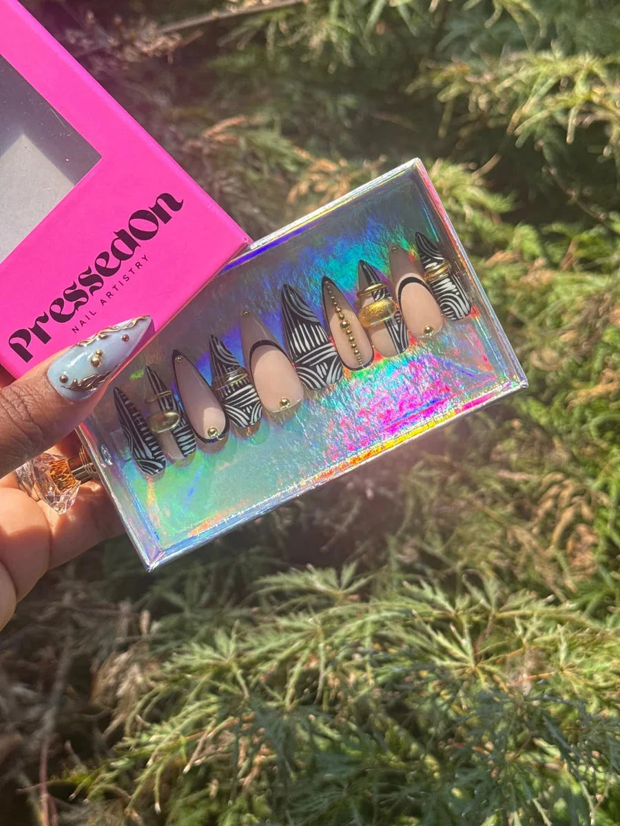 Nude stiletto press-on nails with a black and white zebra stripe pattern. Some nails have gold rings and gold bead details, and one nail is light blue with gold accents.