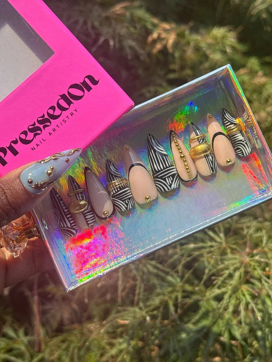 Stiletto press-on nails with a black and white tribal pattern, gold embellishments, nude and light blue accents.