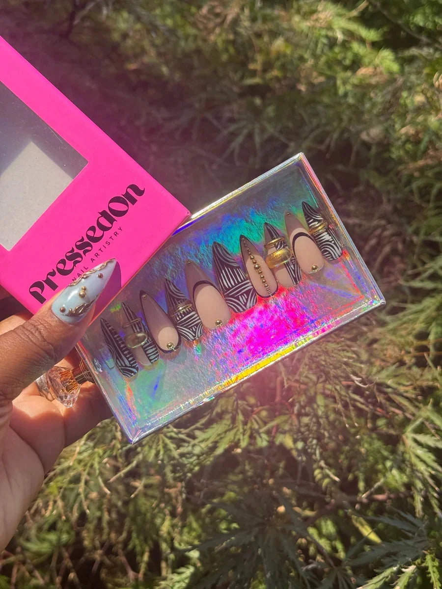 Stiletto press-on nails featuring nude, black and white tribal patterns, and gold embellishments. One nail is light blue with gold accents.