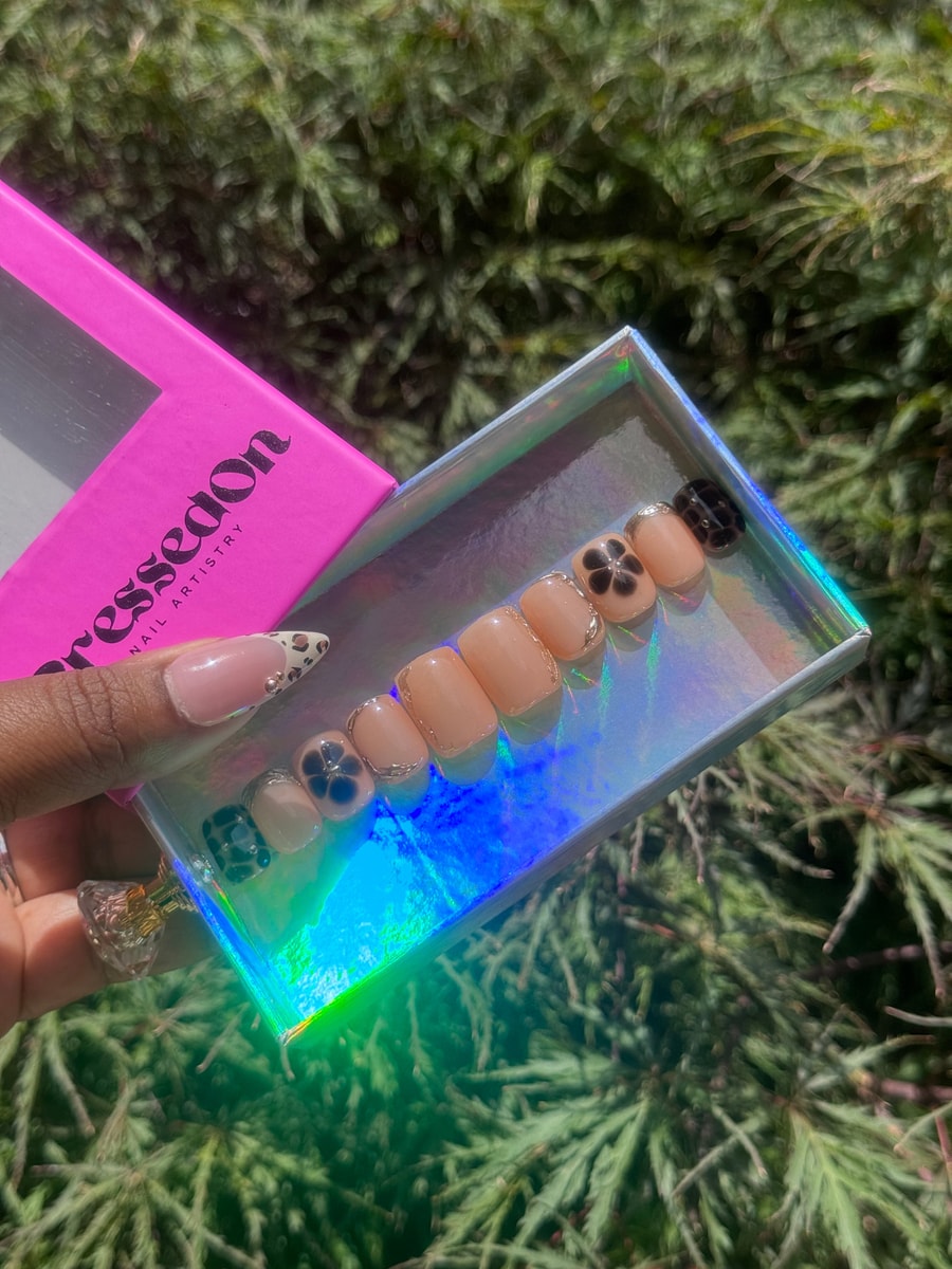 Set of nude, super short press-on nails with subtle black flower designs and gold trim. Some nails feature black and white spotted patterns.