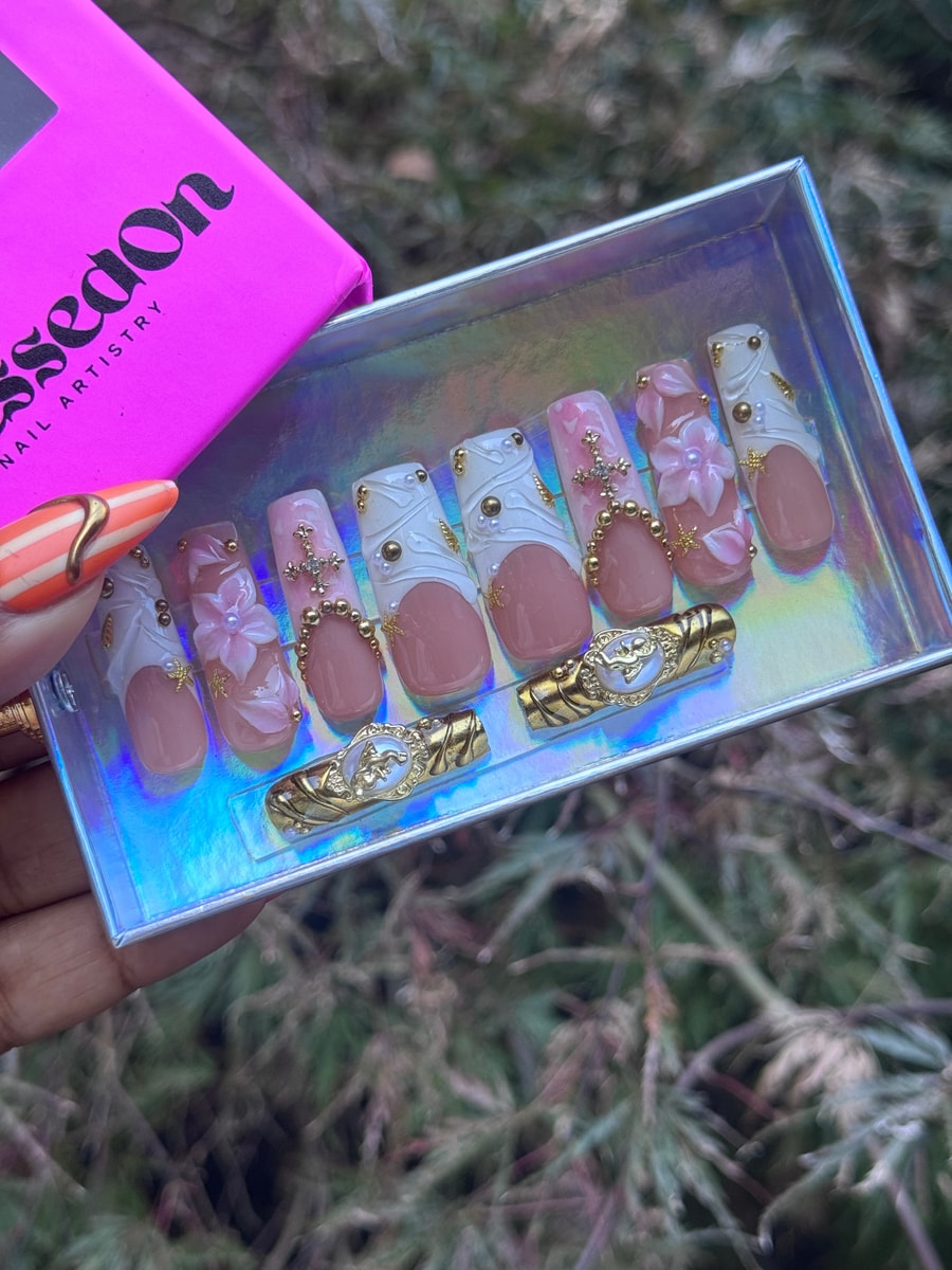 Medium length press-on nails display a pink and white floral design. Gold beads accent the nails, and they feature subtle French tips.