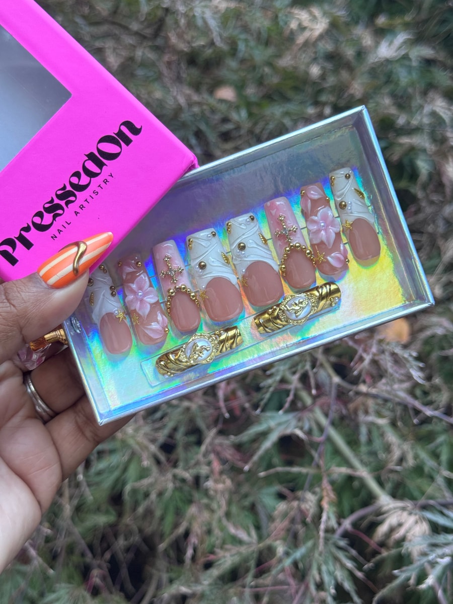 Medium length press-on nails with a nude base, adorned with white swirl designs, gold embellishments, floral accents, and decorative charms.