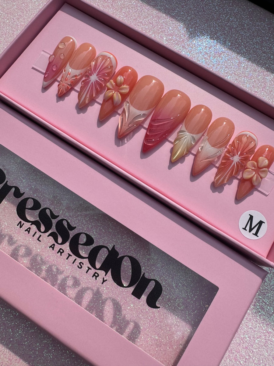 A set of medium-length press-on nails in orange and pink tones, featuring fruit slice designs, floral embellishments, and abstract patterns.