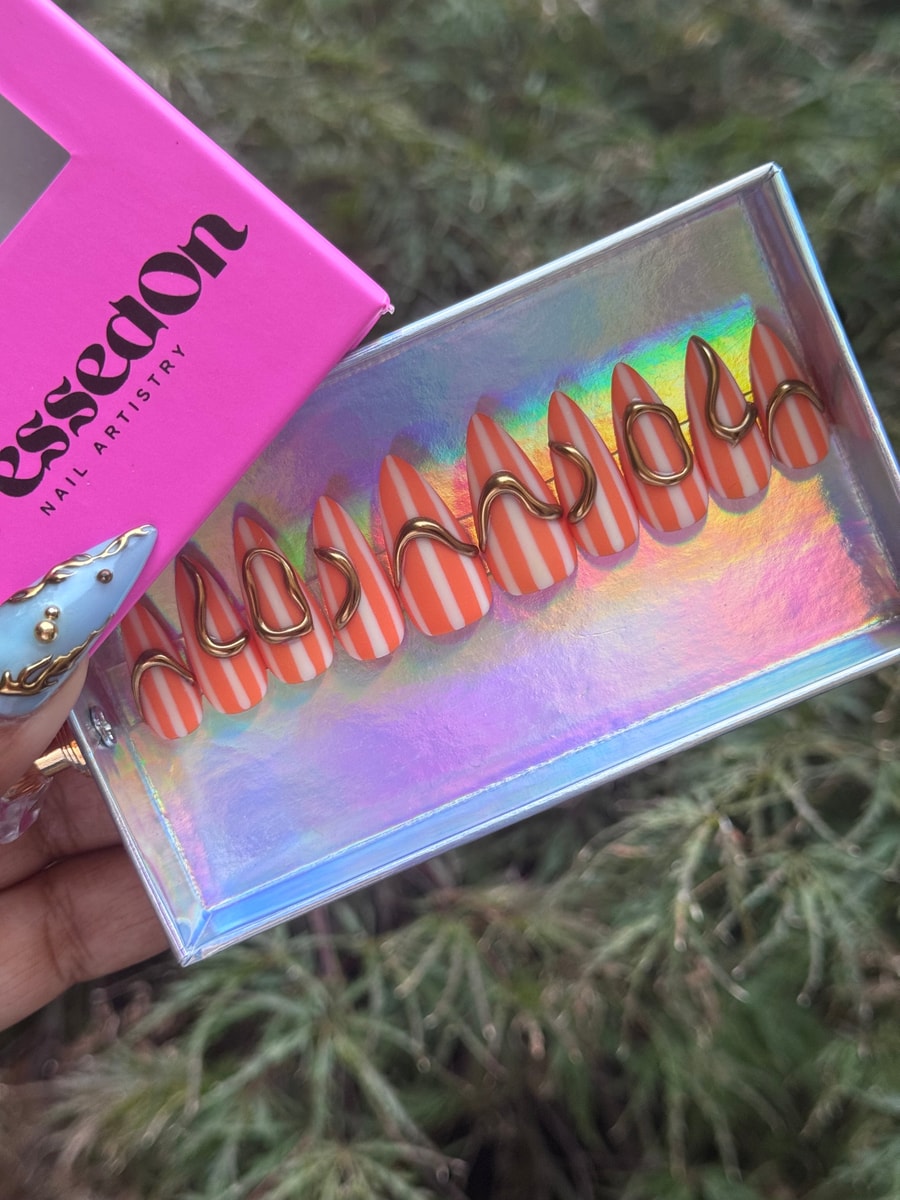 Almond-shaped short press-on nails with orange and white stripes. Gold wire accents embellish each nail.