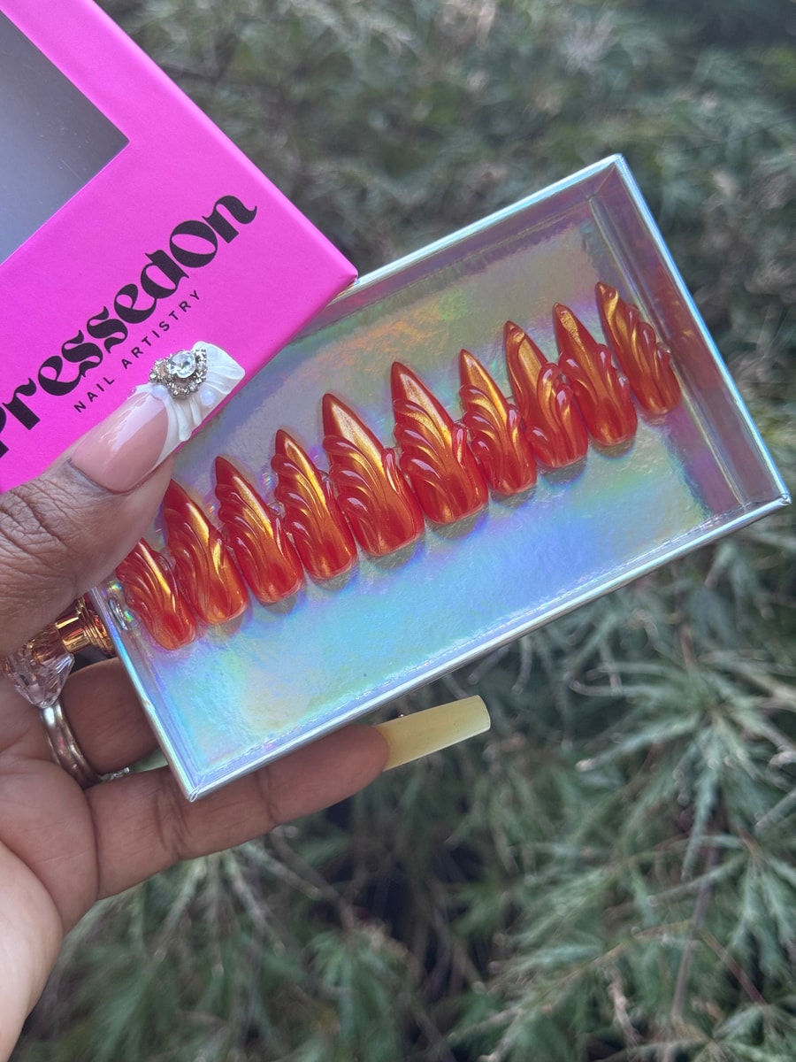 Orange stiletto press-on nails with a glossy finish and a distinctive winged design. These are medium-length nails.