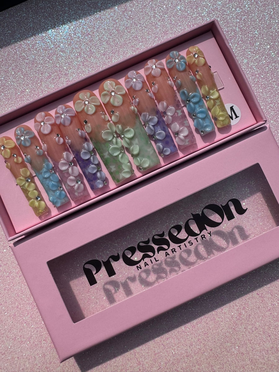 Extra long press-on nails with a pastel gradient and 3D flower designs. The nails feature a mix of pink, blue, purple, and green with glitz accents.