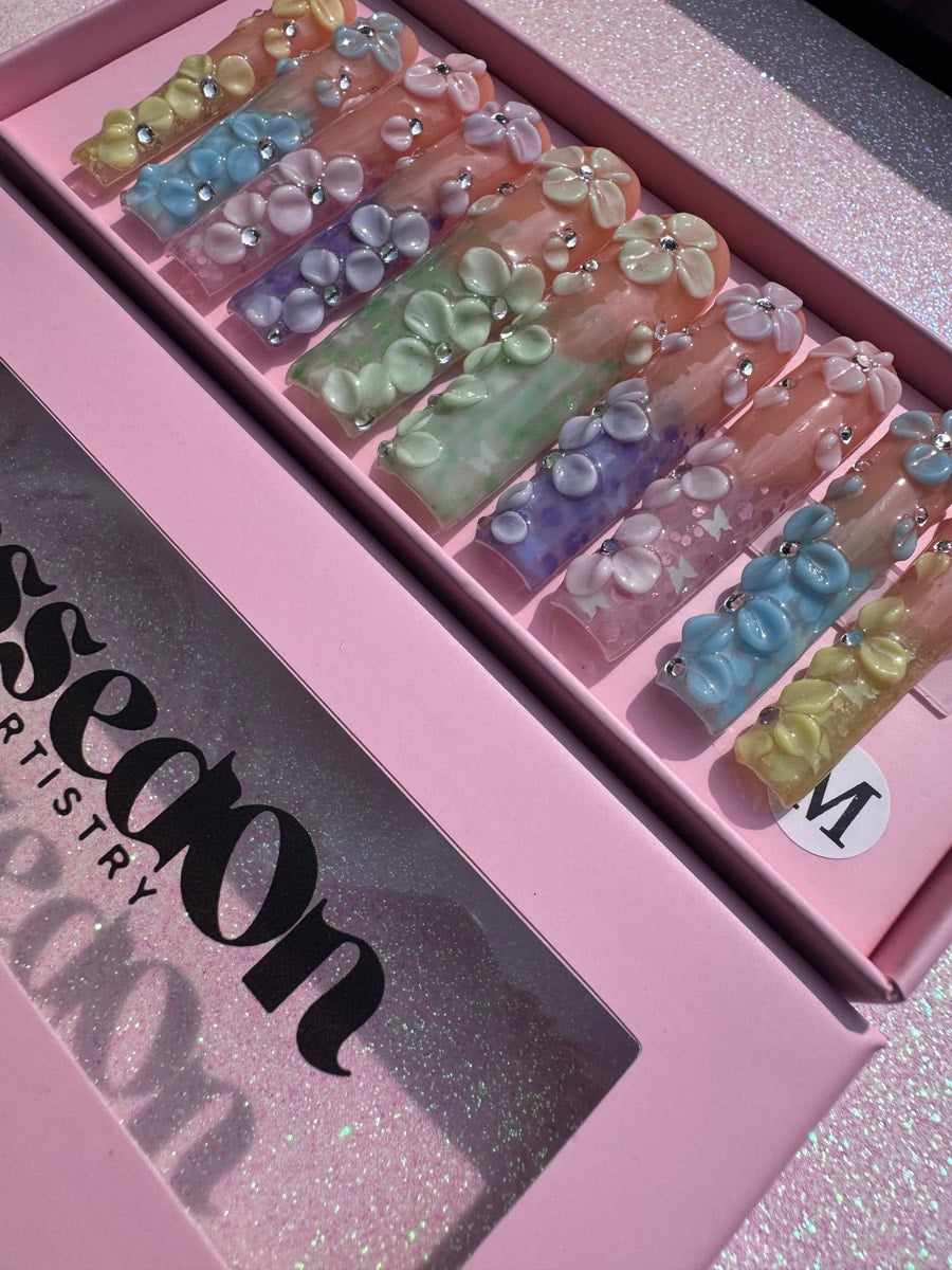 Xtra long press-on nails in pastel colors feature flower charms with crystal centers. The set includes yellow, blue, pink, purple, green, and peach nails.