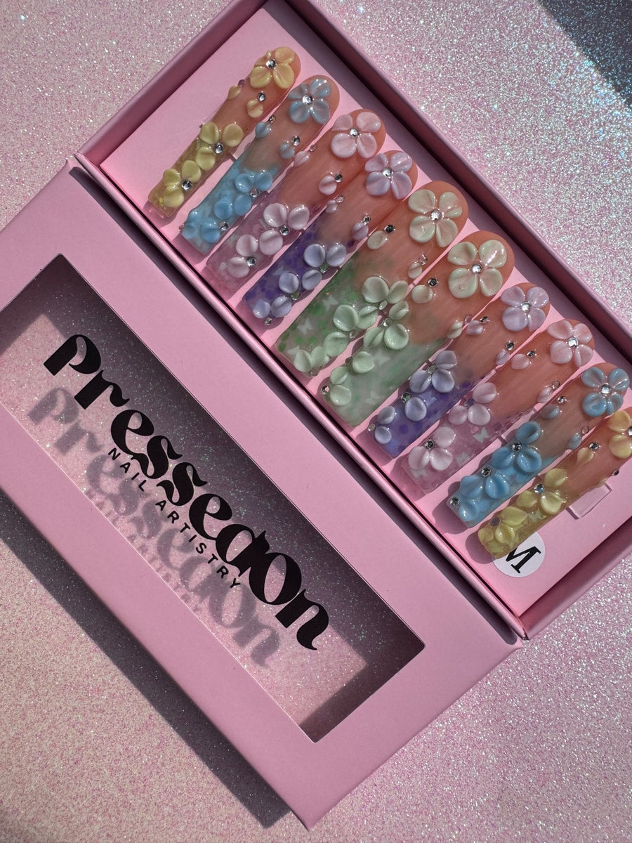 Xtra long press-on nails decorated with pastel yellow, blue, lilac, green, and white flower embellishments. Each flower has a sparkly rhinestone center.