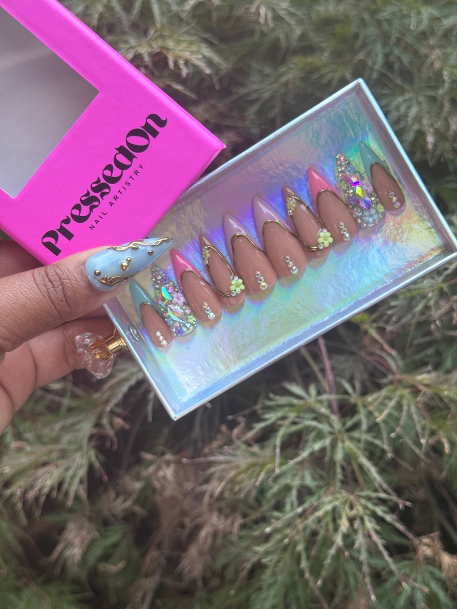 Short stiletto press-on nails feature tan bases and pastel tips, accented with gold outlines, rhinestone details, and small flower charms.