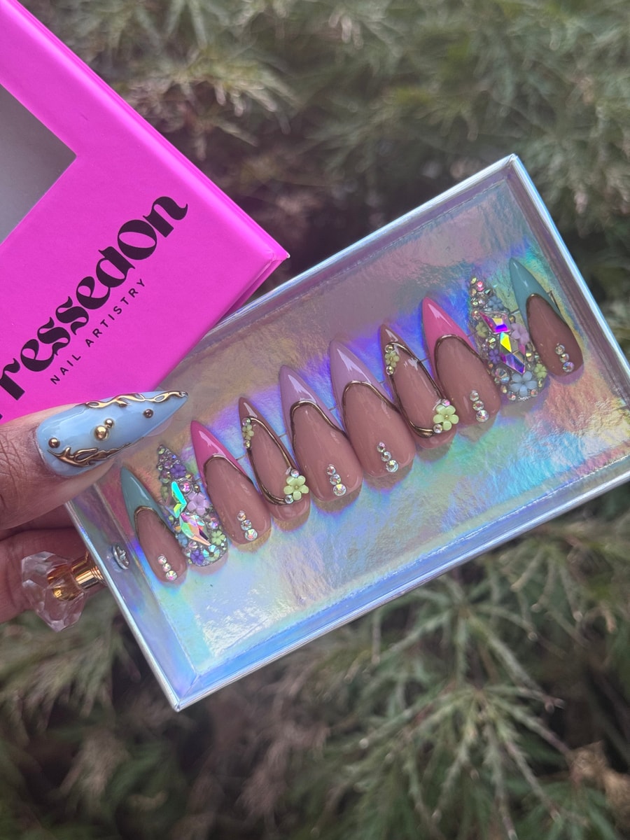 Pastel stiletto press-on nails feature light pink, lavender and blue tips, embellished with gold outlines, flower charms and sparkling rhinestones for a decorative look.