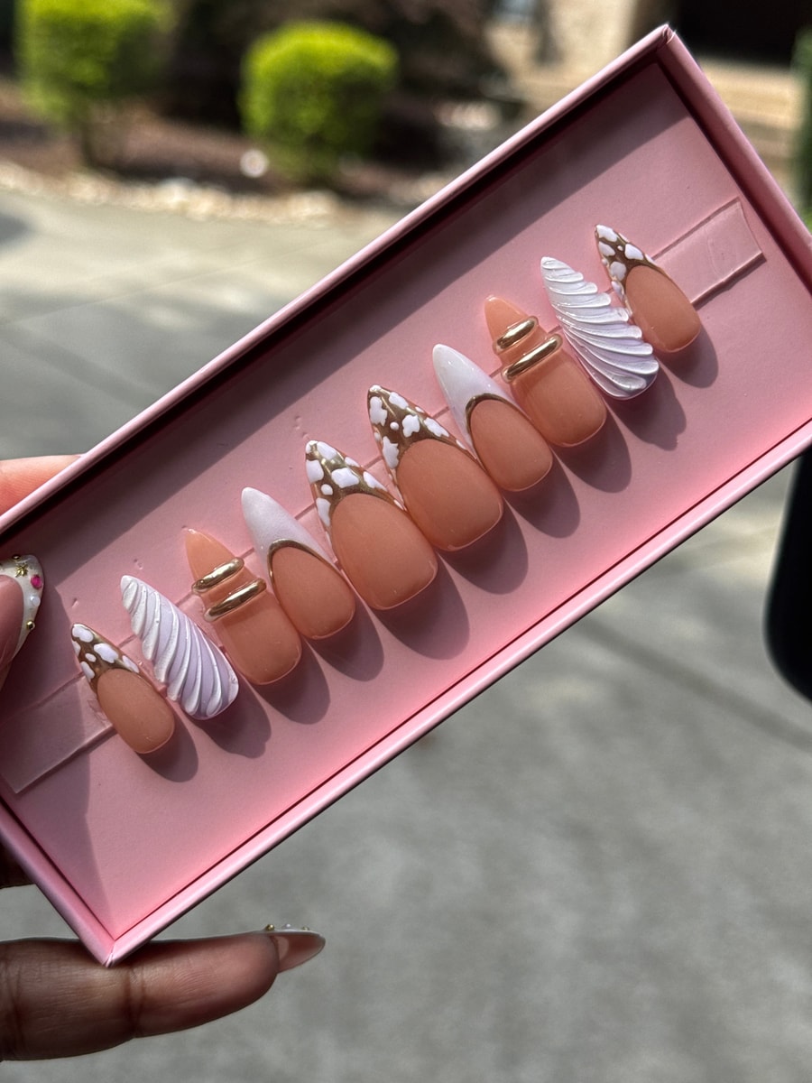 Short peach press-on nails with cloud designs in white and gold, along with chrome accents. Some nails feature a white tip or a gold ring accent.