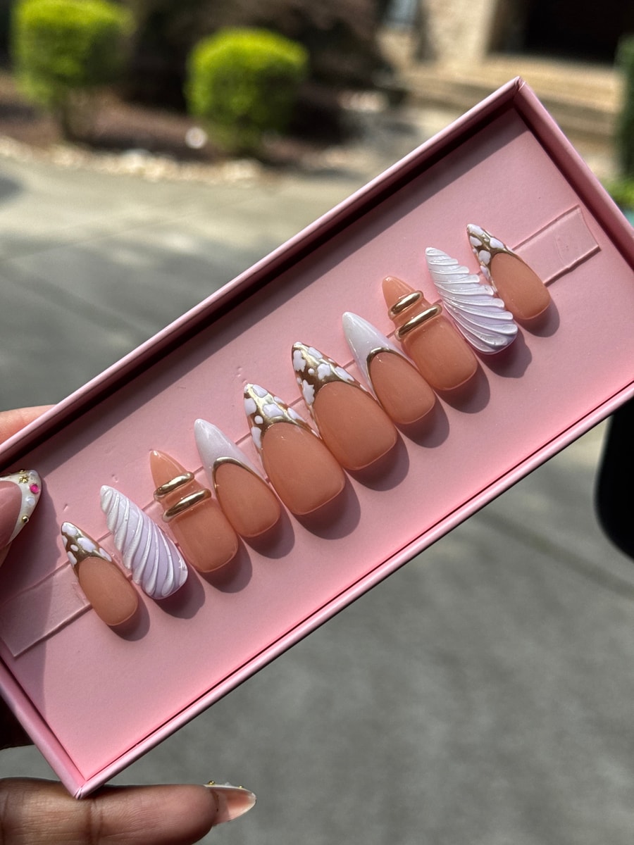 Short peach press-on nails are decorated with white cloud swirls, gold chrome accents, and French tips. A few nails have unique gold ring or foil decorations.