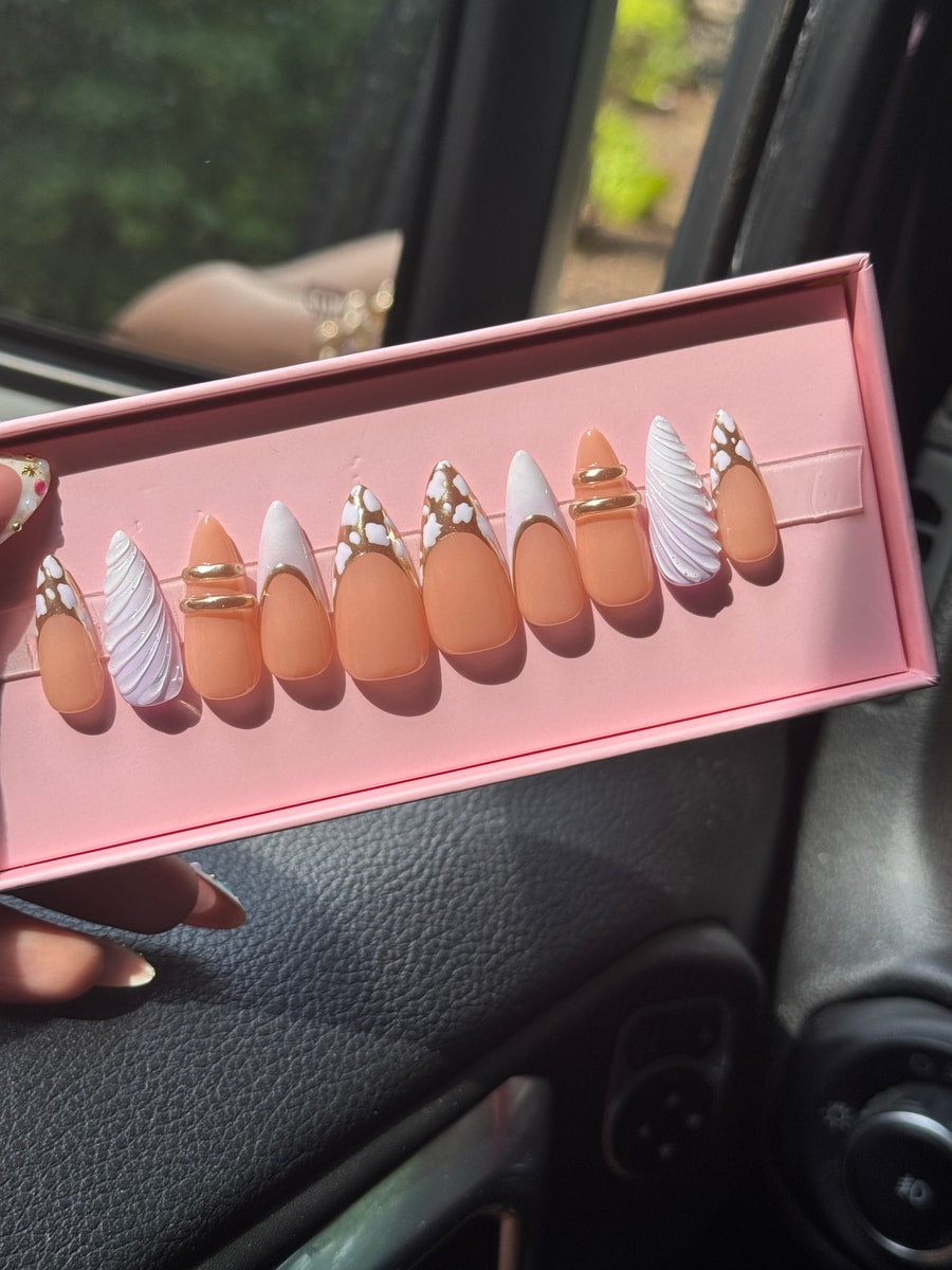 Peach almond-shaped press-on nails with French tips in white and gold chrome, featuring gold ring accents and white swirl designs.