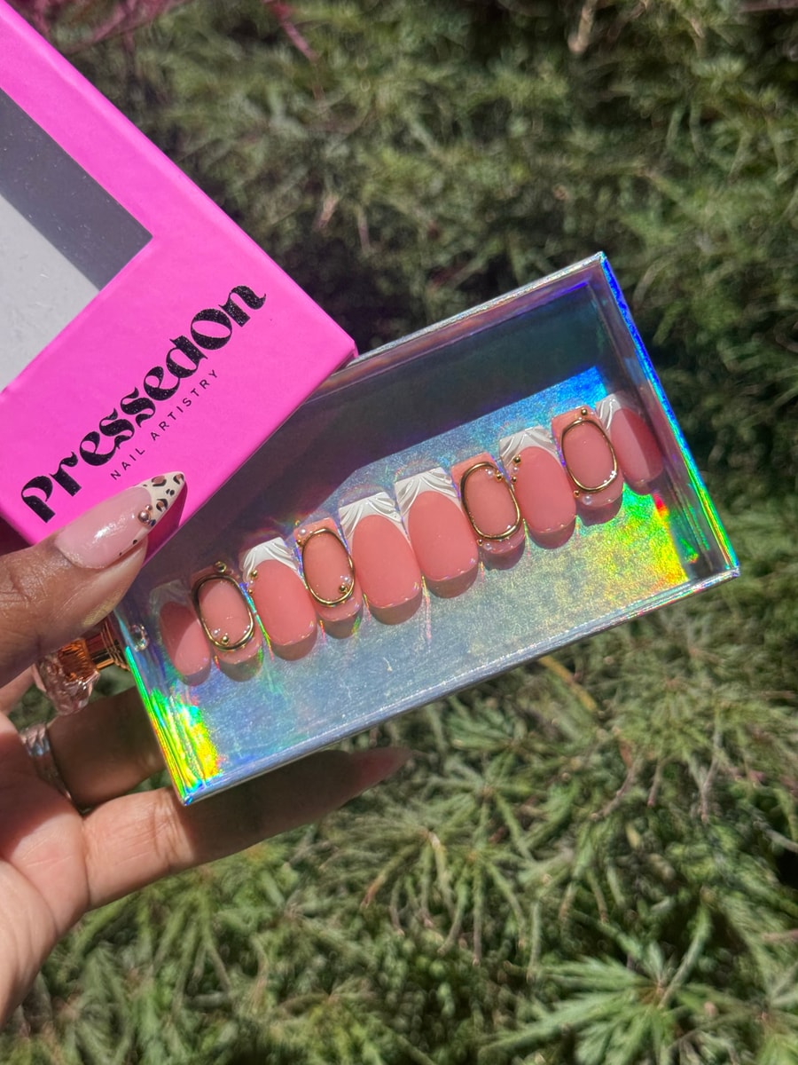 Short peach press-on nails with white French tips, gold oval charms, and pearl accents. A sophisticated and stylish nail design.