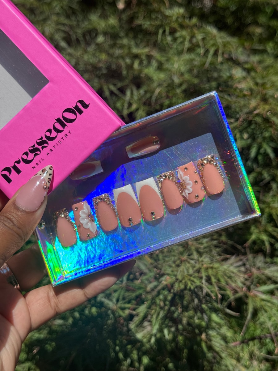 These are peach-colored short press-on nails, some featuring rhinestone accents, a white French tip, a gradient brown French tip, and a white flower charm.