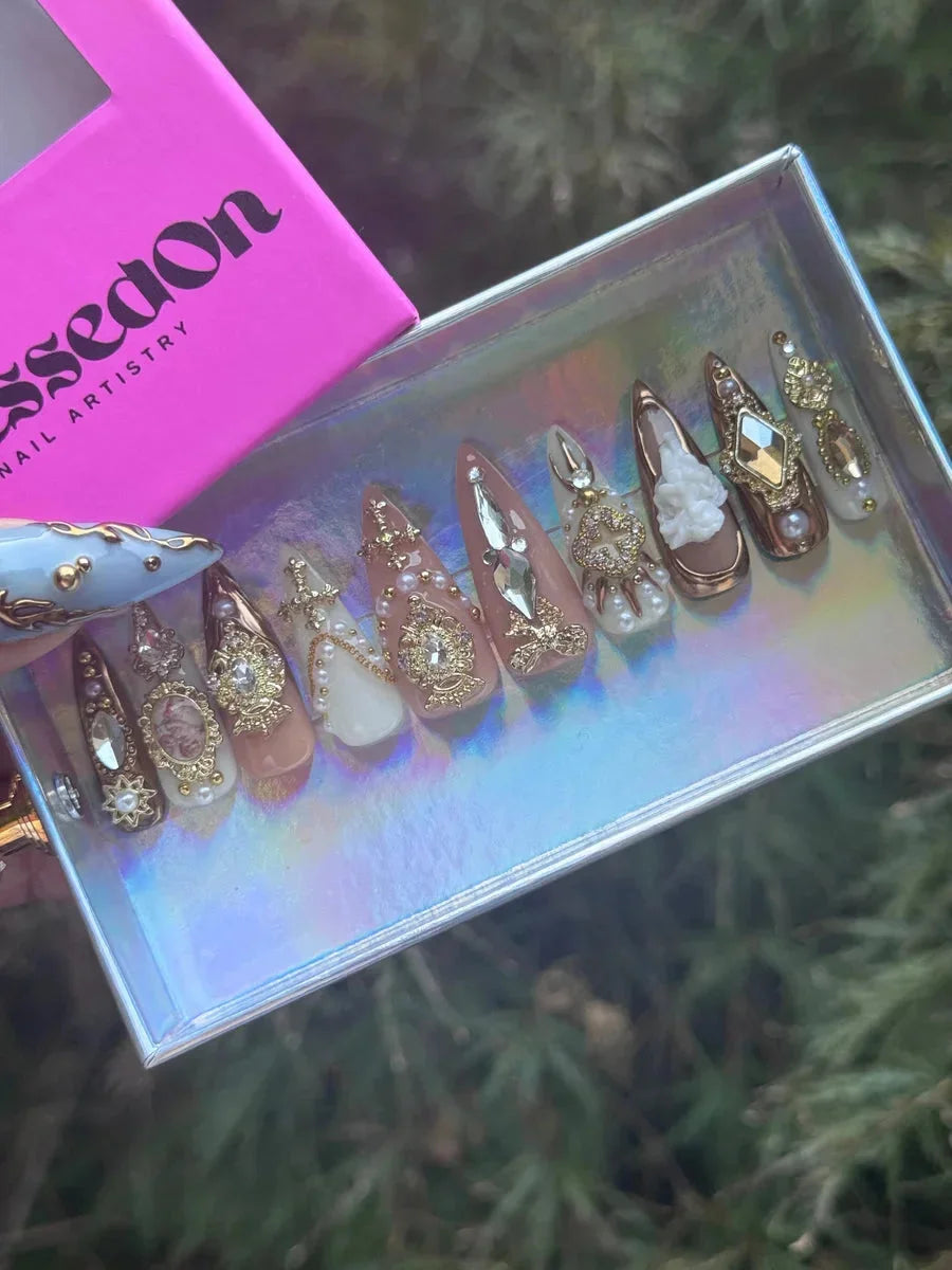 Set of almond-shaped press-on nails with nude, brown, and white colors. These nails are adorned with pearl and rhinestone embellishments, gold accents, and a delicate white floral design.