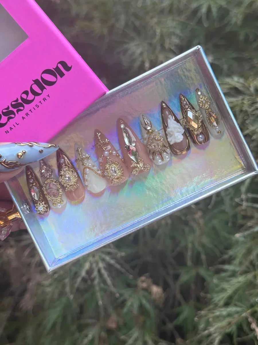 Short almond-shaped press-on nails featuring pearl and rhinestone embellishments, intricate gold detailing, and white floral designs on nude, brown, and white backgrounds.