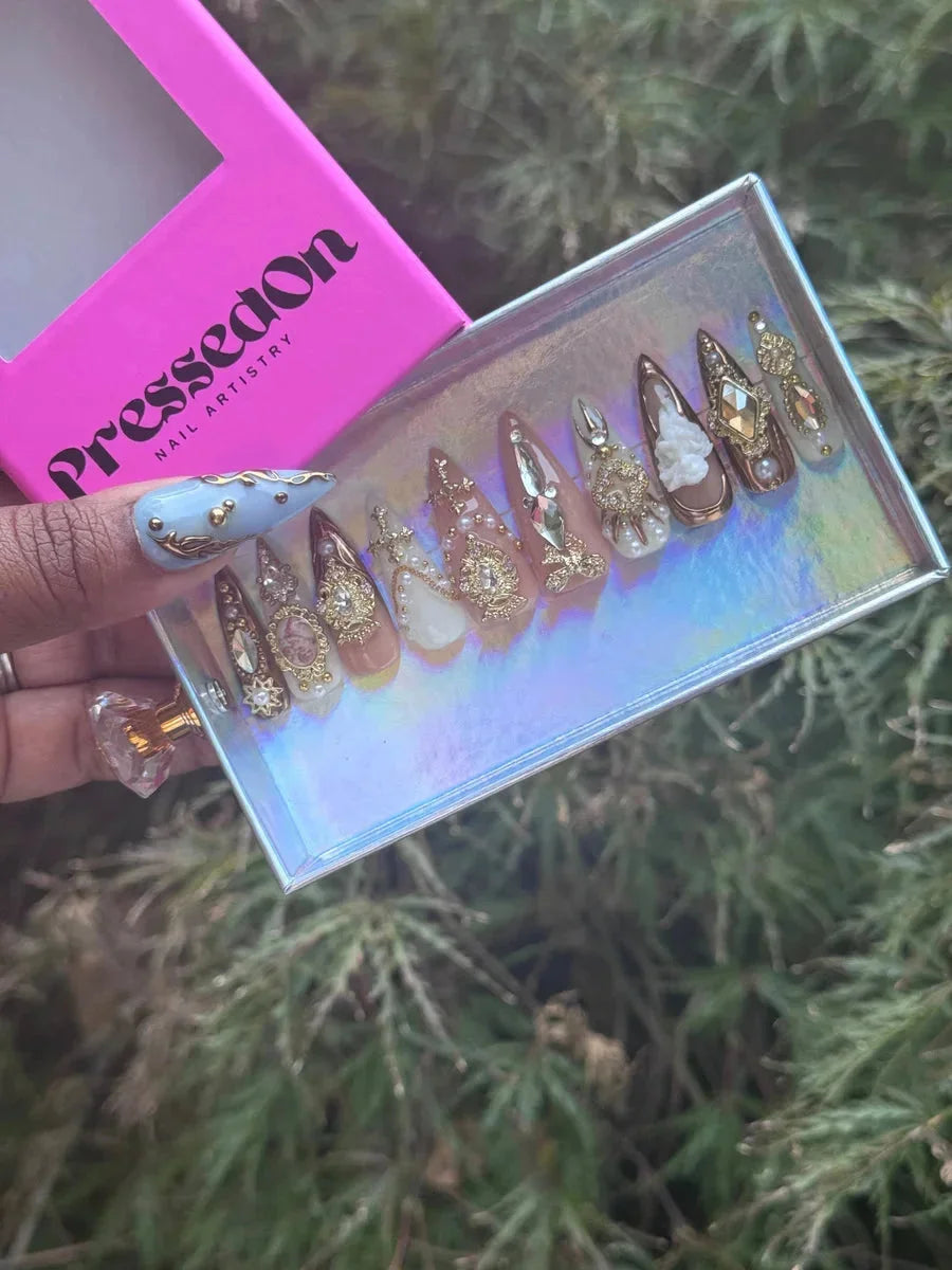 Short almond press-on nails featuring pearl and rhinestone accents, with gold details and white floral designs on nude, brown, and white backgrounds.