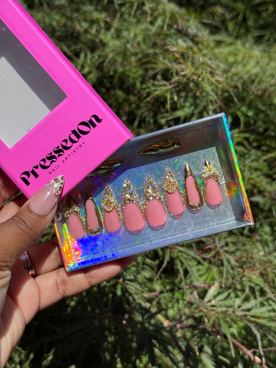 Medium-length pink oval and stiletto press-on nails feature gold edges, rhinestone details, and gold bling charms.