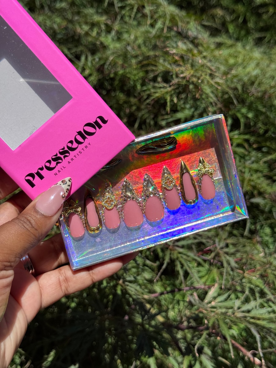 Pink press-on nails with gold edging and rhinestone decorations. Some nails have large rhinestone and gold embellishments.