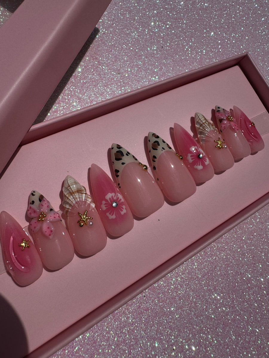 Pink press-on nails featuring a cheetah print design, floral accents, and a seashell embellishment. Each nail is also decorated with rhinestones and small gold beads.