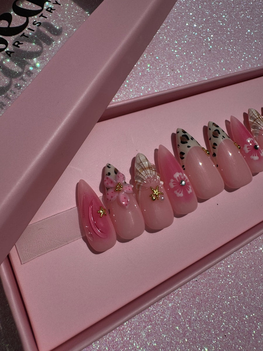 Pink medium-length press-on nails feature cheetah print tips and various 3D decorations including flowers, seashells, and small jewels.