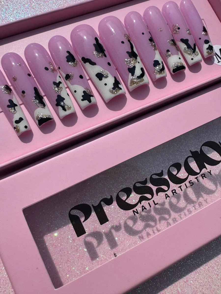Pink press-on nails feature a cow print design with black spots and white patches, accented with silver glitter. The nails are long and coffin-shaped.