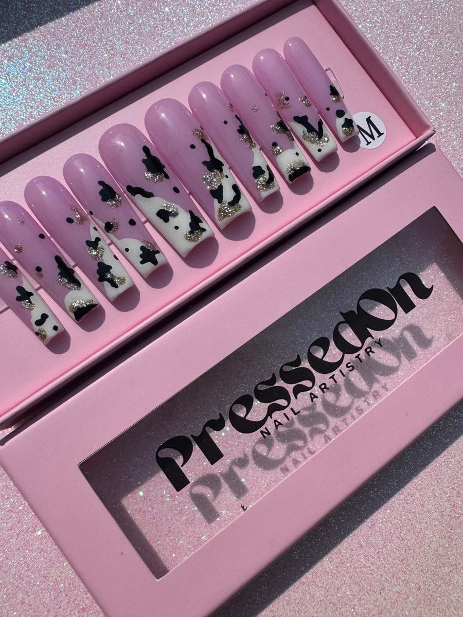 A set of pink press-on nails features a long, coffin shape and a cow print design with white and black spots, accented by silver glitter. Each nail is designed for easy, instant application.
