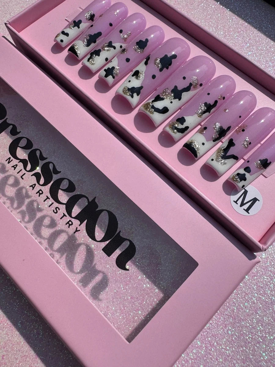 A set of long press-on nails featuring a pink base with black cow print spots. The nails have white tips accented with metallic gold glitter.