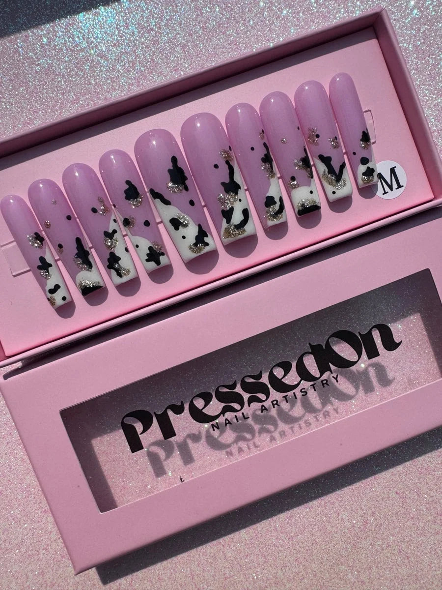 Long, pink press-on nails with a cow print design are shown. Some nails have a white base with black spots, while others feature glitter accents.