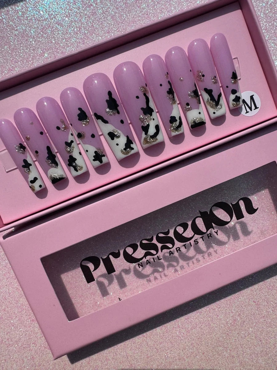 Set of pink, long, coffin-shaped press-on nails with a cow print design and glitter accents. The nails have black spots on a pink base, while others have white tips with silver glitter.