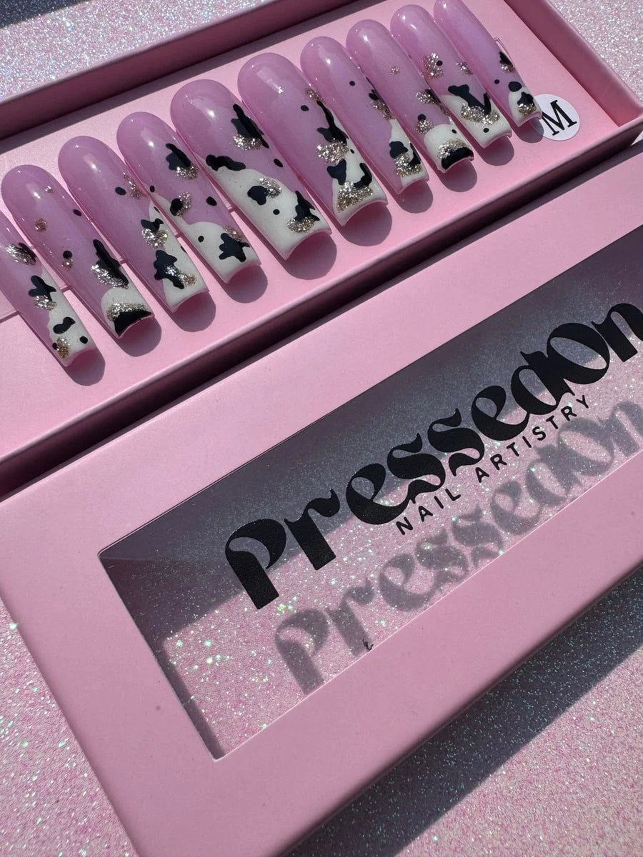 Set of long, pink press-on nails decorated with a cow print design. Nails have black spots, white tips, and silver glitter accents.